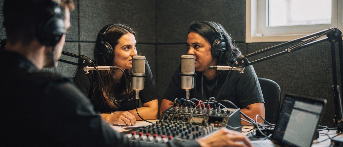 Two presenters in studio headphones speak into condenser microphones in a panelled podcast booth while a sound engineer adjusts levels at a mixing desk