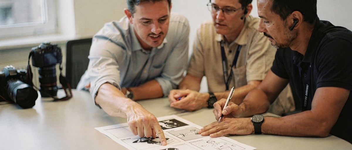 A client points at a printed storyboard on a meeting-room table while two video crew members review the shot list and make notes, a camera body resting beside them