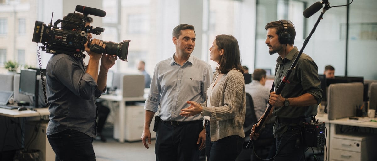 A film crew with a camera and boom microphone films two colleagues talking as they walk through a bright open-plan office
