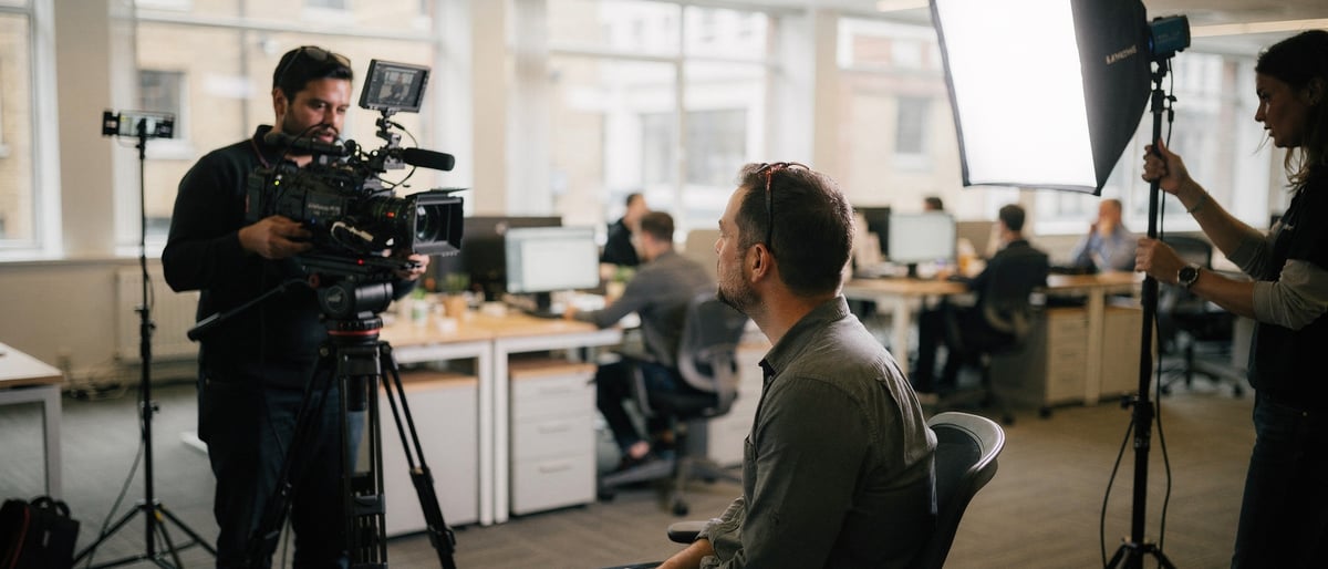 A two-person film crew records a video interview with an employee in a bright open-plan office, one operator behind a cinema camera on a tripod and another adjusting a softbox light while colleagues work at desks behind them
