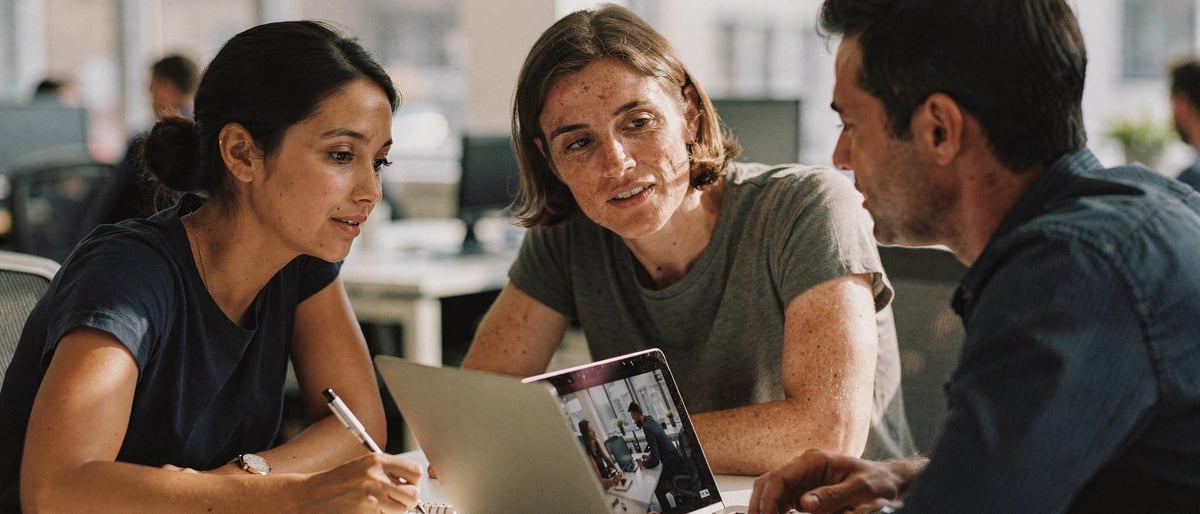 Two new staff members and a manager watch an internal training video on a laptop at a desk in a bright office