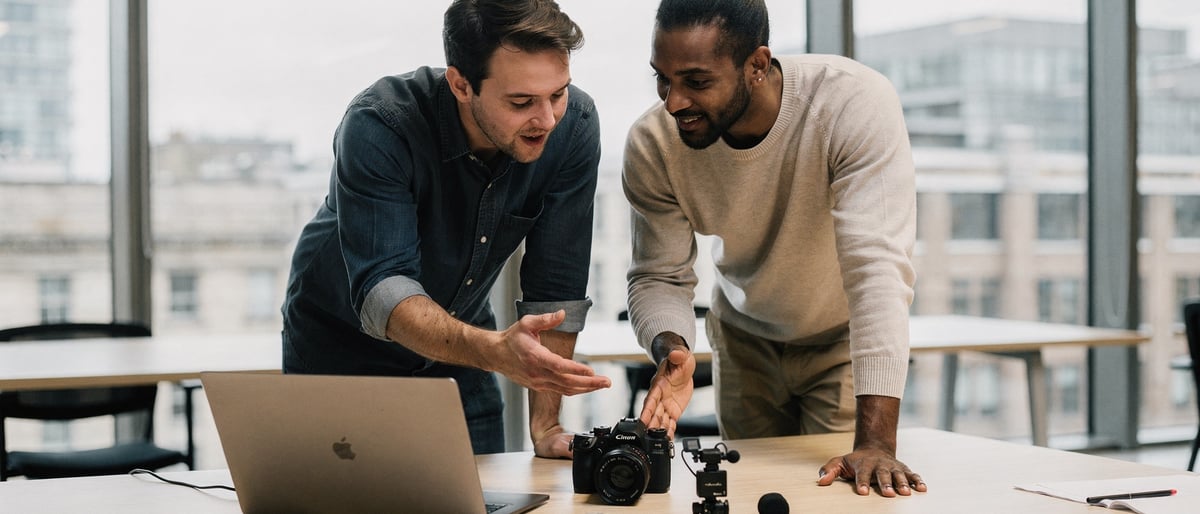 Two video producers in a bright London office comparing a small camera and a clip-on microphone on a laptop as they plan a shoot