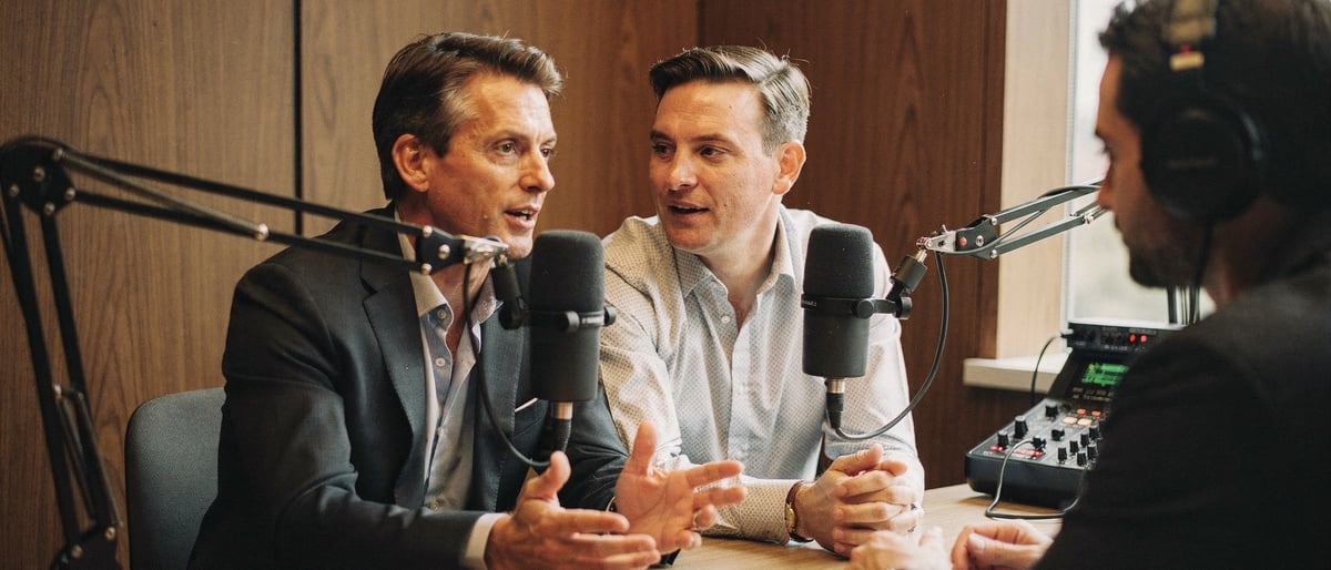 Two executives talking into broadcast microphones in a wood-panelled podcast booth while a producer monitors audio levels on a mixer