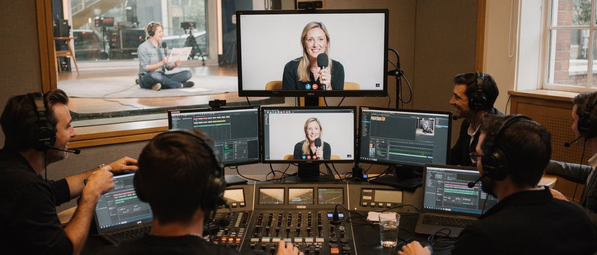 A production crew in a London studio gallery watching a remote guest on a video-call monitor while a presenter waits on the studio floor