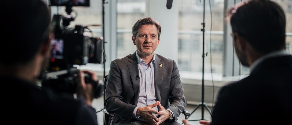 A composed executive being filmed mid-interview in a bright London studio, camera operator in the foreground