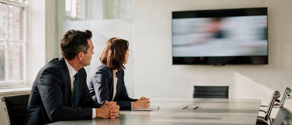 Two executives in a bright London meeting room watching a short brand film on a wall-mounted screen during a client pitch