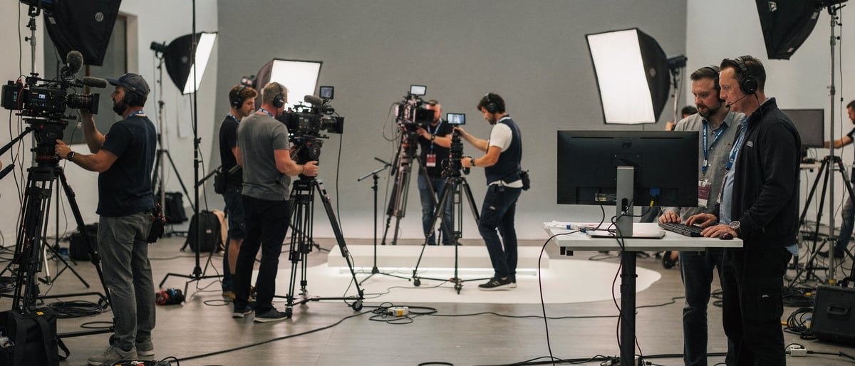 A film crew rigging cameras and lighting around a studio set while a director and producer review a monitor during a fast production build