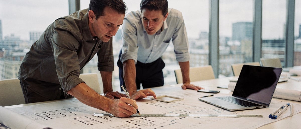 Two architects and a project manager in a bright London office reviewing printed building plans and a laptop together