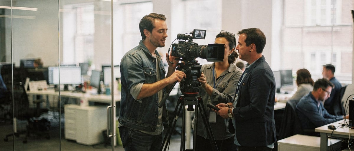 A presenter and a two-person film crew set up a camera on a tripod in a bright modern London production studio, with an office floor visible through a glass partition