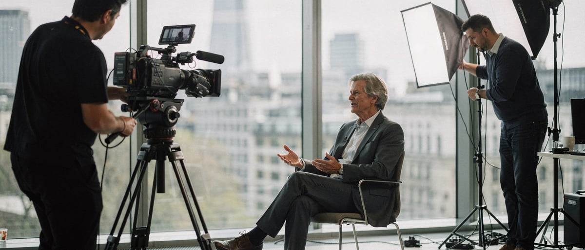 A two-person film crew shooting a seated business executive in a bright modern office, one operating a cinema camera on a tripod while the other adjusts a softbox light