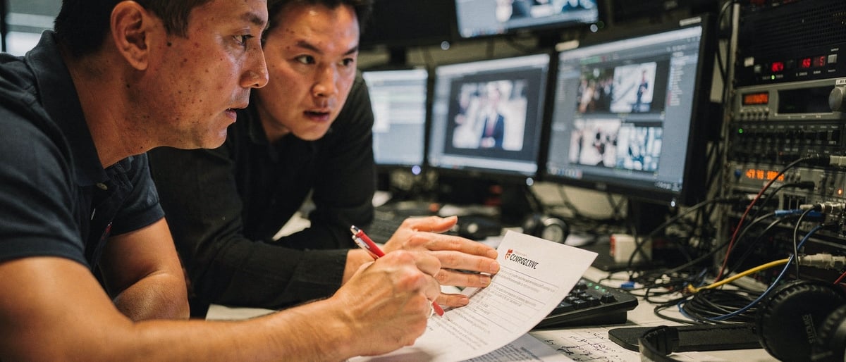 Two colleagues in an edit suite reviewing footage on monitors and marking up a printed script