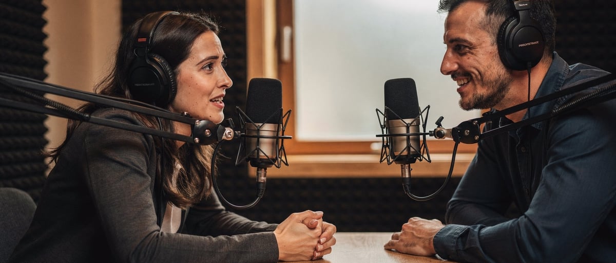 A nervous first-time guest in a podcast booth listens as a friendly producer reassures her before recording, both wearing headphones at a small table with studio microphones.
