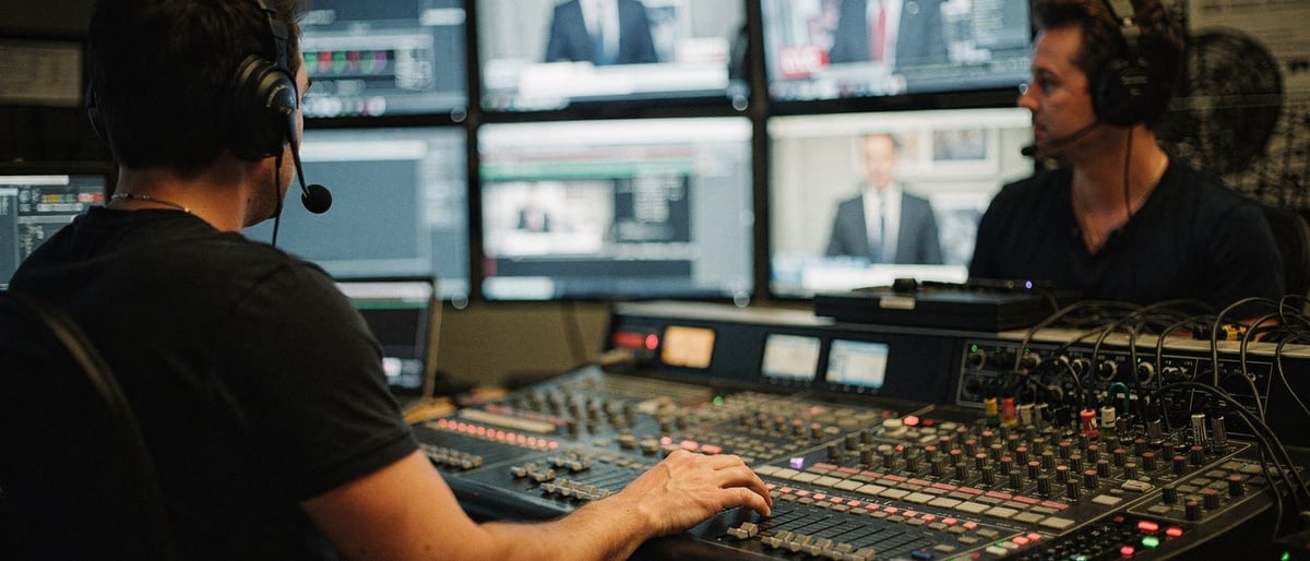 Two production crew members in a broadcast control gallery watching a live presenter feed across vision-mixing desks and monitors, one adjusting a fader