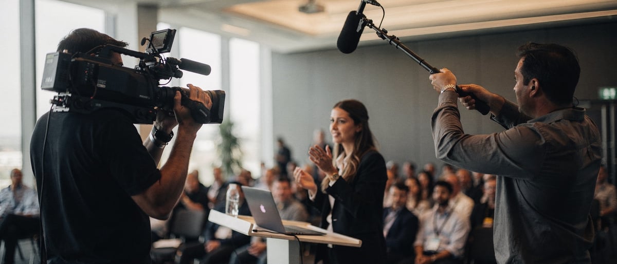 A video crew films a businesswoman giving a keynote on a conference stage in front of a seated audience