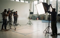 A film crew sets up cameras, tripods and lighting around a presenter's mark on a London studio floor as a technician adjusts a softbox