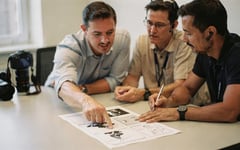 A client points at a printed storyboard on a meeting-room table while two video crew members review the shot list and make notes, a camera body resting beside them