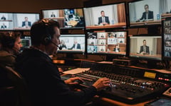 Production team in a dim control gallery watching a live multi-camera panel discussion on the monitor wall as a vision mixer cuts between speakers at the desk