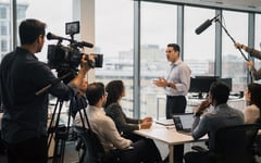 A film crew records a presenter delivering a training session to staff in a bright modern office, with a camera operator and a colleague holding a boom microphone