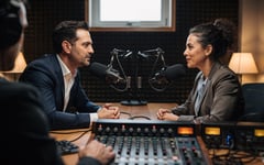 Two colleagues being recorded for an interview in a sound-treated booth with studio microphones on boom arms while an audio engineer adjusts a mixing console