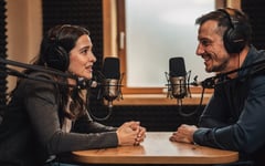 A nervous first-time guest in a podcast booth listens as a friendly producer reassures her before recording, both wearing headphones at a small table with studio microphones.