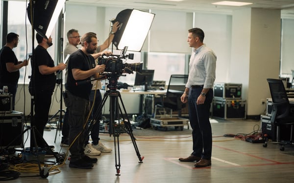 A film crew resetting lights and a tripod-mounted camera between takes as a presenter waits on a marked spot in a production space