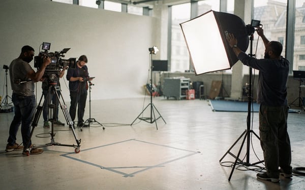 A film crew sets up cameras, tripods and lighting around a presenter's mark on a London studio floor as a technician adjusts a softbox