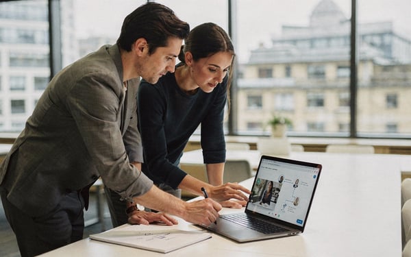 Two marketing colleagues in a bright London office clicking through an interactive video on a laptop while sketching a customer journey on a notepad