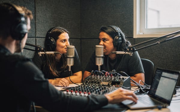 Two presenters in studio headphones speak into condenser microphones in a panelled podcast booth while a sound engineer adjusts levels at a mixing desk