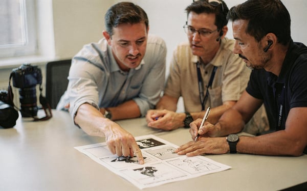 A client points at a printed storyboard on a meeting-room table while two video crew members review the shot list and make notes, a camera body resting beside them