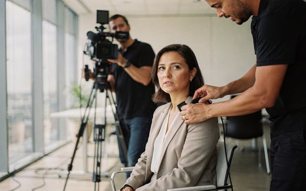 A film crew sets up a client testimonial interview in a bright office, a sound recordist clips a lapel mic onto a seated businesswoman while a camera operator frames the shot