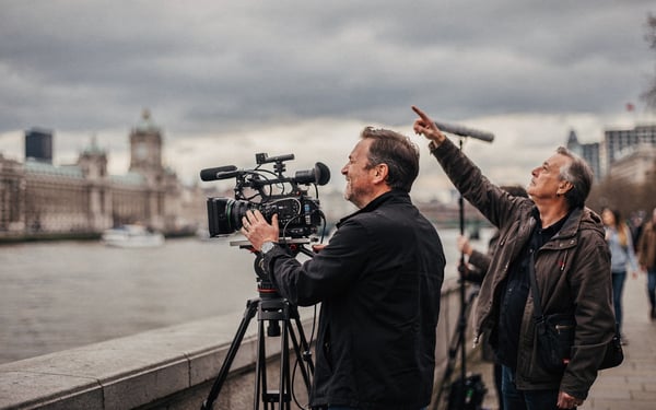 A documentary film crew filming on the South Bank promenade beside the River Thames under an overcast sky