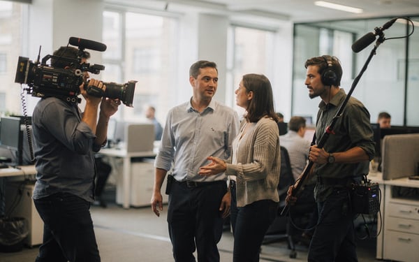 A film crew with a camera and boom microphone films two colleagues talking as they walk through a bright open-plan office