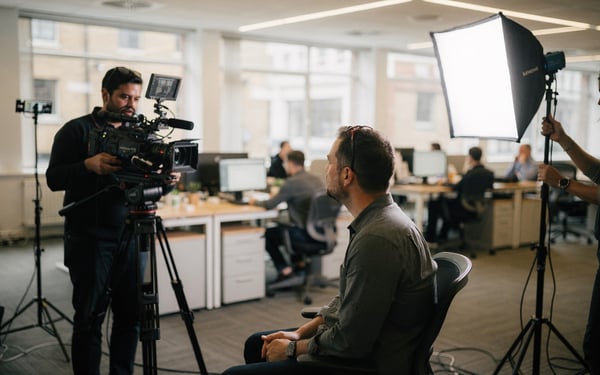 A two-person film crew records a video interview with an employee in a bright open-plan office, one operator behind a cinema camera on a tripod and another adjusting a softbox light while colleagues work at desks behind them