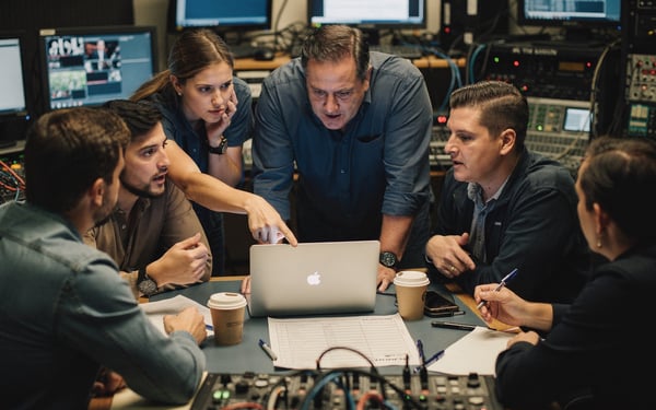 A production crew gathered around vision-mixing desks in a control gallery, replanning a shoot at short notice over a laptop and shot list