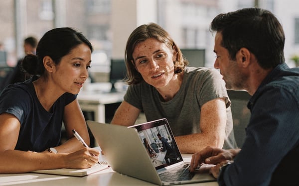 Two new staff members and a manager watch an internal training video on a laptop at a desk in a bright office