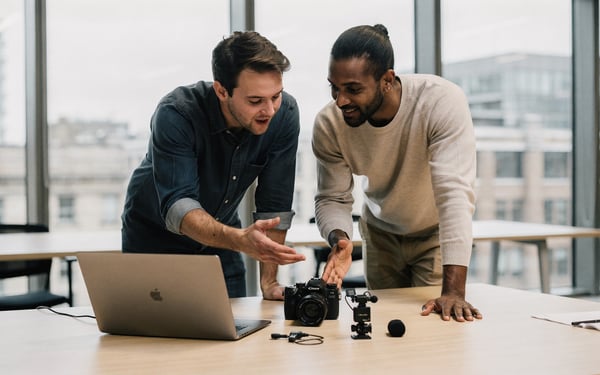 Two video producers in a bright London office comparing a small camera and a clip-on microphone on a laptop as they plan a shoot