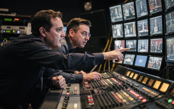 Two broadcast engineers at a vision-mixing desk in a dim control gallery switching to a backup feed during a live show