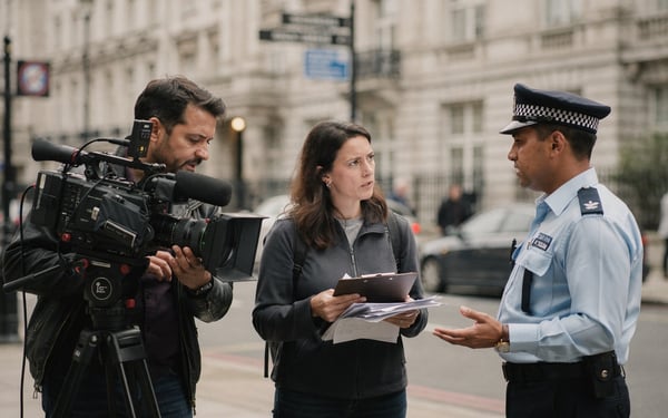 A small film crew with a tripod-mounted camera pause on a London street corner as a council officer speaks to the producer holding a clipboard
