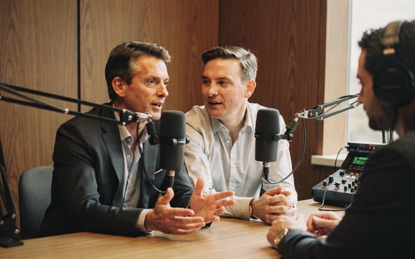 Two executives talking into broadcast microphones in a wood-panelled podcast booth while a producer monitors audio levels on a mixer