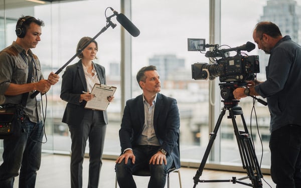 A film crew filming a business interview in a London office, with a camera on a tripod, a boom microphone and a producer checking a shot list