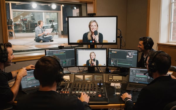 A production crew in a London studio gallery watching a remote guest on a video-call monitor while a presenter waits on the studio floor