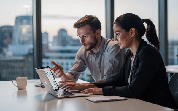 Two colleagues in a bright London office reviewing overnight analytics results on a laptop early in the morning, one pointing at the screen