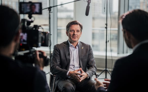 A composed executive being filmed mid-interview in a bright London studio, camera operator in the foreground