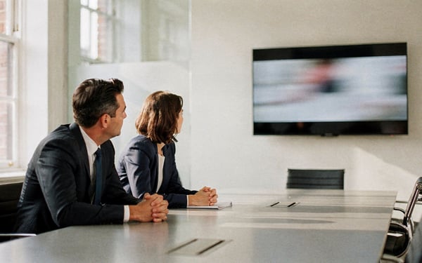 Two executives in a bright London meeting room watching a short brand film on a wall-mounted screen during a client pitch