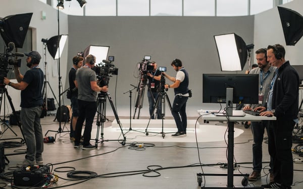 A film crew rigging cameras and lighting around a studio set while a director and producer review a monitor during a fast production build