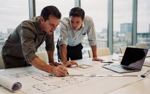 Two architects and a project manager in a bright London office reviewing printed building plans and a laptop together