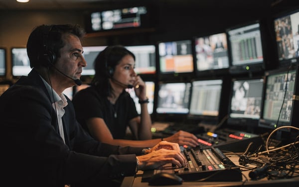 A broadcast director and vision mixer watching a bank of preview monitors in a dim television control gallery during a live show