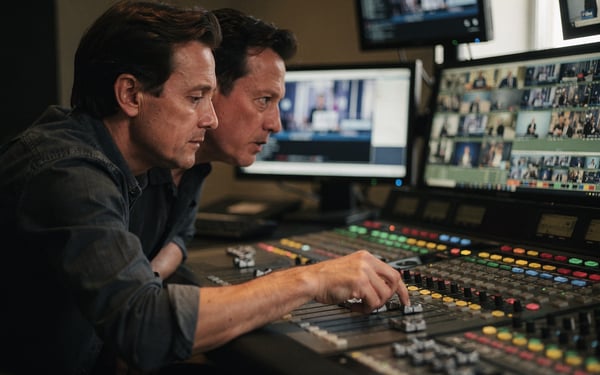 Two broadcast operators at a control gallery desk comparing a live programme feed across vision-mixing panels and monitors