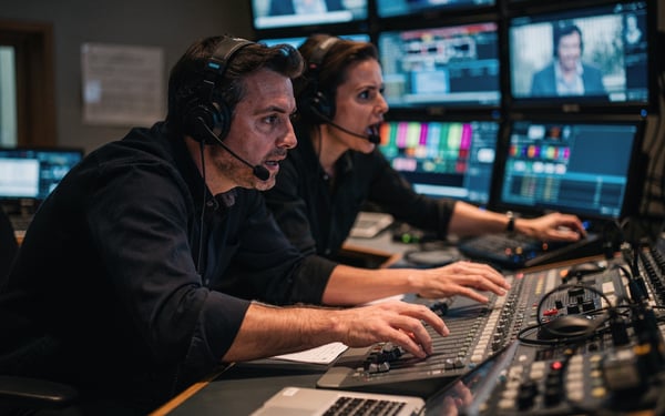 A broadcast director and vision mixer at a control gallery desk watching a wall of monitors during a live show
