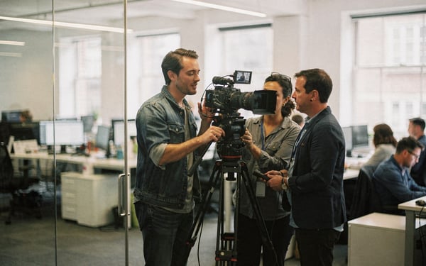 A presenter and a two-person film crew set up a camera on a tripod in a bright modern London production studio, with an office floor visible through a glass partition